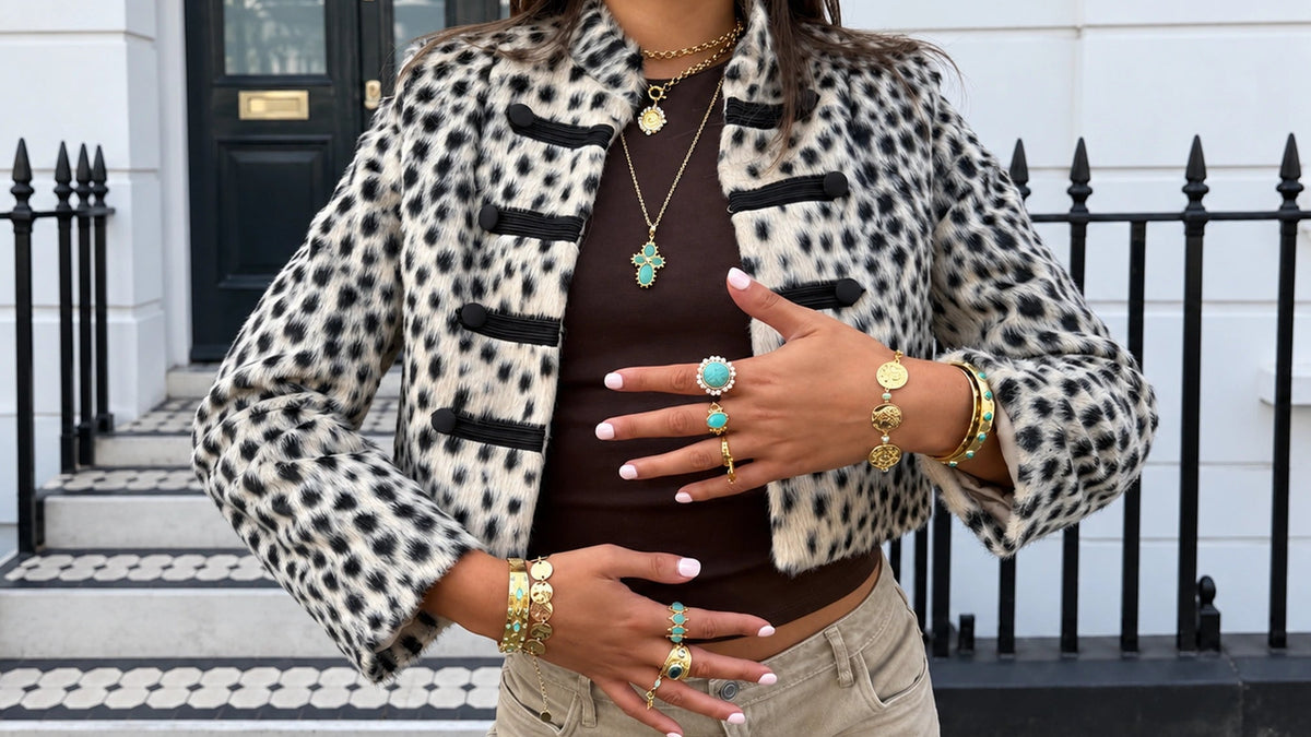 A woman wearing a cropped black-and-white spotted jacket over a dark brown top and beige pants, styled with gold jewelry, standing in front of a white building with a black iron fence.
